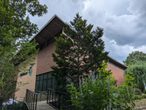 A photograph of the gardens on the UUFR property with the UUFR building. A large deciduous tree is in front of the UUFR brick building, with a pathway on the left side with black guardrails guiding the way from the gardens.